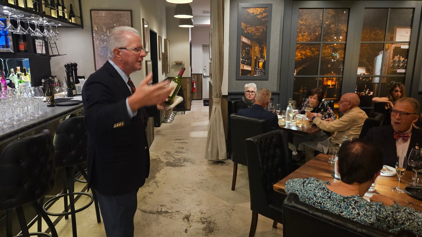 A host in a navy blazer holding a bottle and speaking to the seated dining room at Enology Wine Bar and Bistro