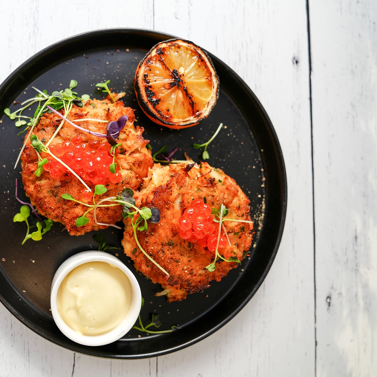 Crab cakes with salmon roe and micro greens on a black plate with charred lemon and aioli