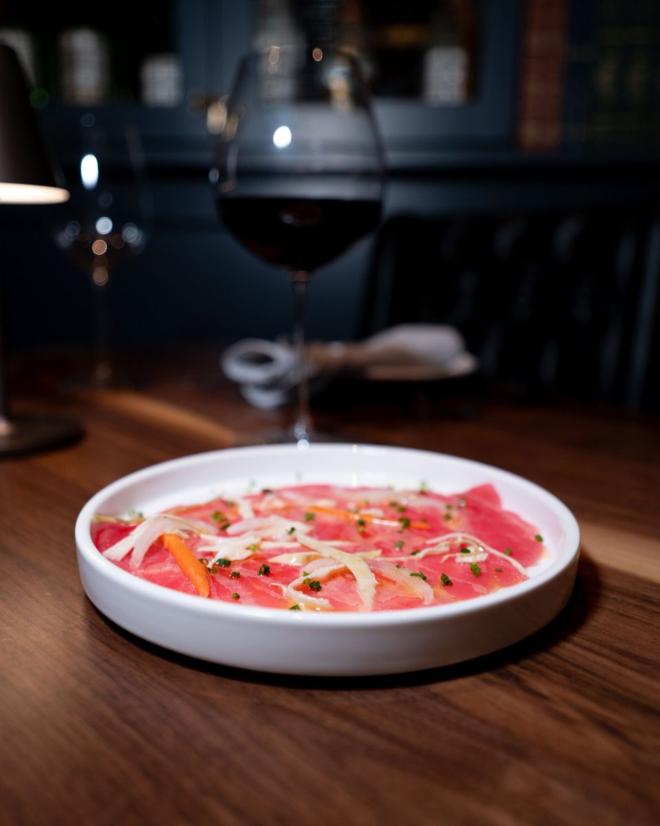 Thinly sliced fish crudo with shaved fennel, pickled carrot, and chives, beside a glass of red wine
