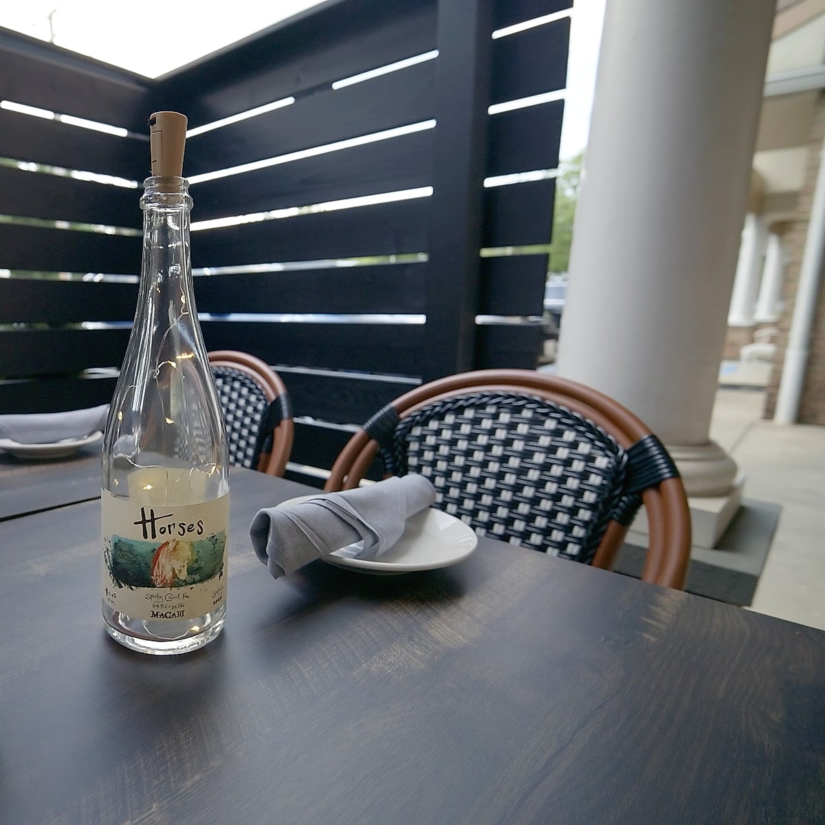 A patio table set with linen and a bottle of white wine, beside bistro chairs and a slatted privacy screen at Enology